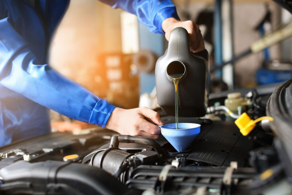 A mechanic in a blue uniform pouring motor oil into a car engine using a funnel, with the engine components and a blurred workshop environment in the background.
