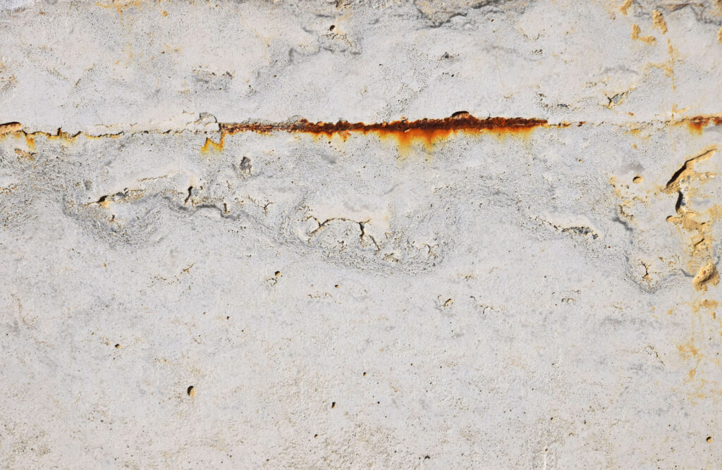 Detailed texture of a concrete wall with a horizontal line of rust indicating metal corrosion beneath, surrounded by patches of peeling paint and surface erosion.