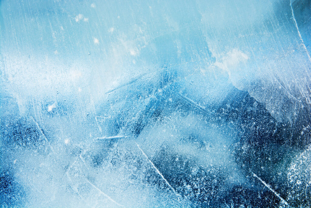 Close-up view of a textured ice surface with intricate patterns of cracks and bubbles, showcasing a range of blue hues from light to dark demonstrating how freezing moisture reacts.