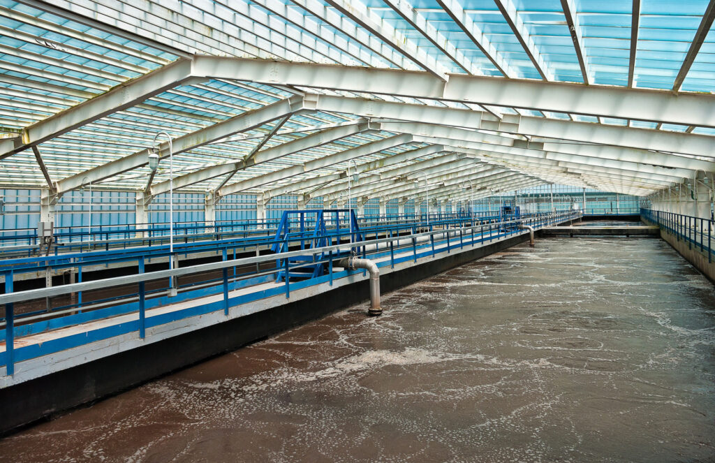 Interior of a large sewage water treatment facility with aeration tanks partially covered by a translucent roof, allowing daylight to filter through. The framework consists of metal beams and glass panels, with blue railings lining the walkway above the frothy, churning water below, indicative of active water purification processes.