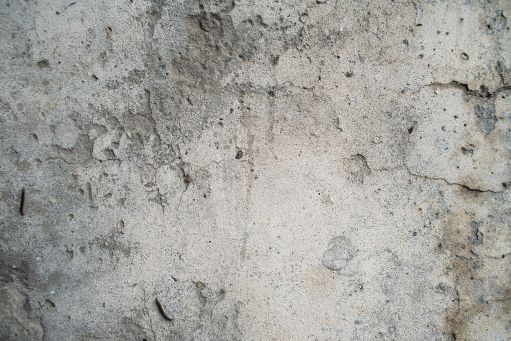 Detailed texture of a concrete wall with natural variations, showing pitting, small cracks, and various imperfections caused by abrasion. The image captures the raw and rugged character of the concrete surface.