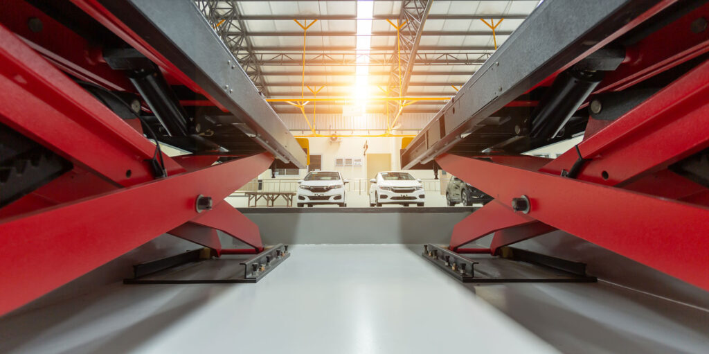 Low angle view inside a modern automated parking garage with a vehicle lift system. The red mechanical structures support cars on elevated platforms, optimizing space utilization. Sunlight filters through the building, casting a warm glow over the steel and concrete interior.