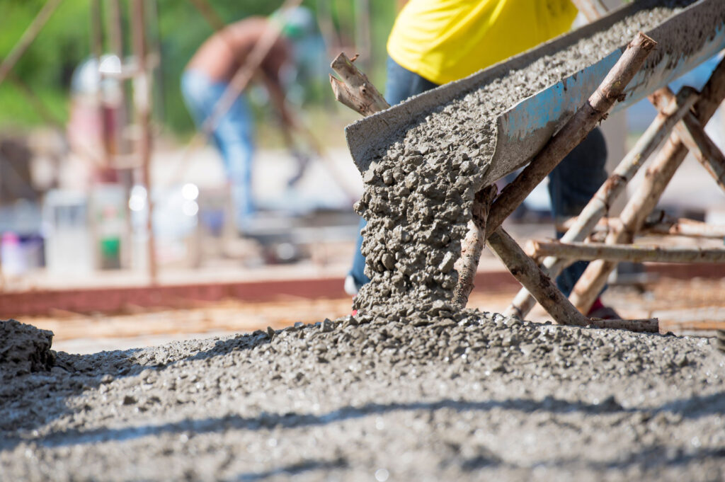 Close-up image showing fresh concrete being poured from a wheelbarrow on a construction site, with a blurred background of workers. The focus on the flowing concrete emphasizes the process of laying foundations.