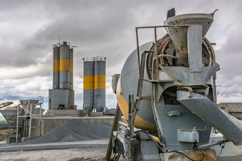 An industrial cement mixer at a construction site with silos in the background under a cloudy sky. The focus is on the weathered and functional machinery, indicative of an active concrete production facility.