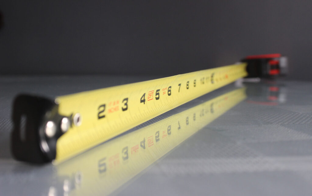 A close-up of a yellow measuring tape extending across a reflective surface, with the focus on the numerical indicators. The shallow depth of field highlights the numbers on the tape, suggesting precision in measurement.