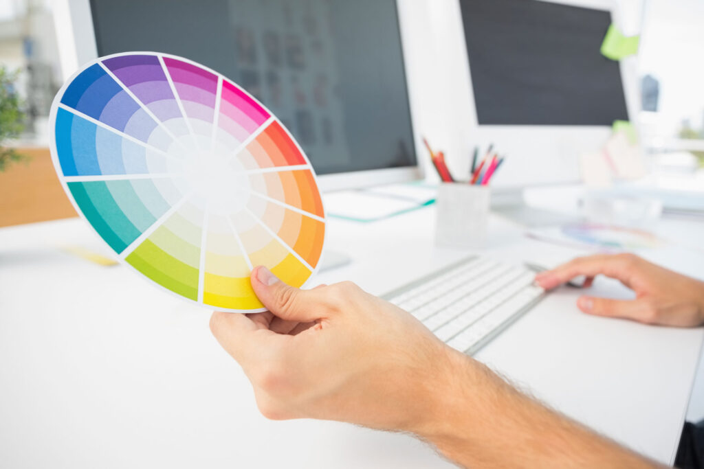 A person's hands are shown holding a color wheel while working on a computer, illustrating a design process in a bright, modern office. The color wheel is in focus, displaying a spectrum of colors, indicating color selection for a creative project.