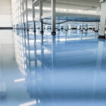 The interior of a vast empty warehouse with a highly reflective epoxy floor that mirrors the white columns and ceiling infrastructure. The blue tint of the space suggests a cool, clean environment suitable for industrial or commercial use. An example of urethane Coatings.