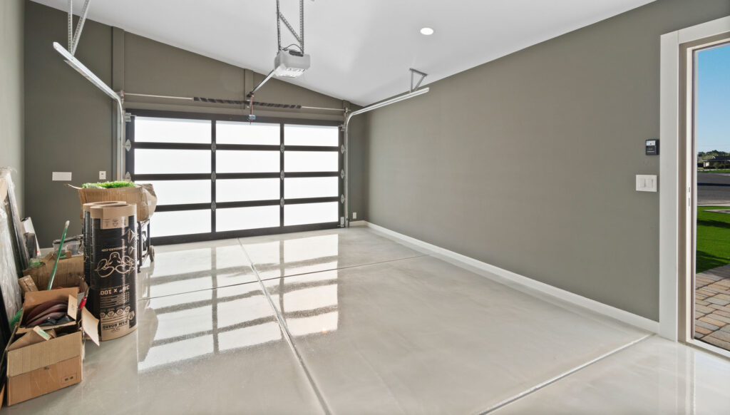 Modern garage interior with a freshly coated epoxy floor reflecting the natural light coming through the large frosted glass door. The walls are painted in a neutral gray tone, and various items, including gardening supplies and storage boxes, are neatly organized to the side. An open door on the right leads to a bright outdoor area.