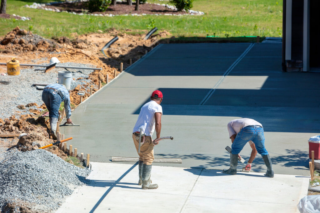 Three construction workers engaged in laying and smoothing new concrete for a new driveway. The workers are using trowels and other tools, focusing intently on creating an even surface. Construction materials and the earthy construction site surround the area being worked on.