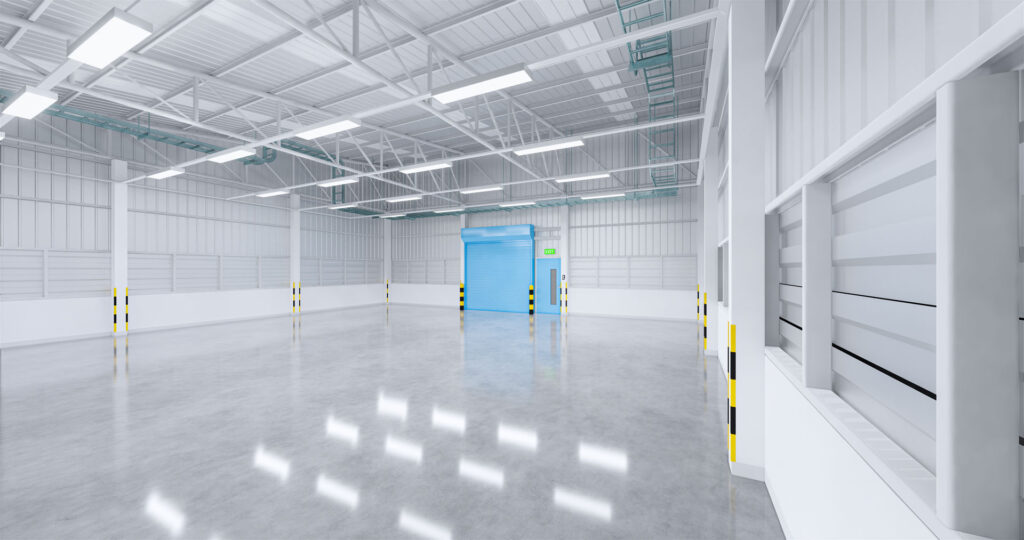 Empty industrial warehouse interior with a polished concrete floor, white walls, and a high ceiling with skylights allowing natural light to create patterns on the floor. The space is equipped with metal infrastructure and features a prominent blue roll-up door marked as an exit, indicating readiness for logistics and storage operations.
