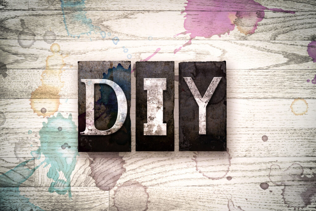 The acronym 'DIY' for 'Do It Yourself' composed of grungy metal letterpress type on a rustic wooden background splattered with colorful paint drips, symbolizing creativity and handcrafted projects.