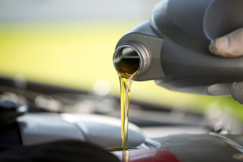 A close-up view of motor oil being poured from a container into a car engine, with a focus on the golden stream of oil. The blurred background suggests this is taking place in a well-lit, outdoor environment.