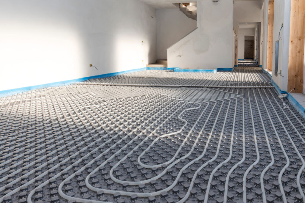 Underfloor heating installation in progress with a network of white pipes laid out in a serpentine pattern on top of insulation panels. The construction scene shows unfinished walls and protective blue tape along the edges, suggesting ongoing interior work with a plan to cover the radiant heating system with polished concrete or concrete epoxy coatings.
