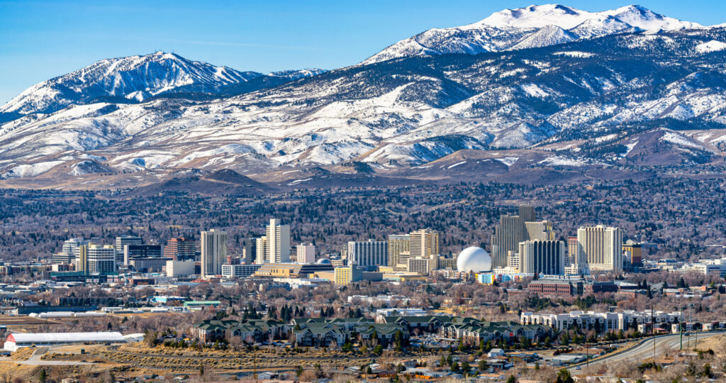 Panoramic view of a cityscape with a mixture of residential and commercial buildings against the backdrop of majestic snow-capped mountains and clear blue skies. The city's architecture includes high-rise buildings and a distinctive white dome structure, integrated seamlessly into the natural landscape.