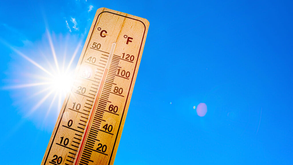 A wooden thermometer scales up into a clear blue sky with the bright sun gleaming from behind, suggesting high temperatures. The thermometer shows both Celsius and Fahrenheit measurements, emphasizing the heat of a sunny day which can effect concrete surface temperature.