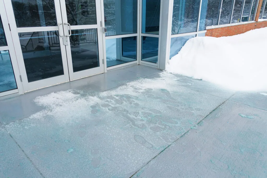 A glass double door entrance covered with a thin layer of winter snow and ice, with visible salt spread to melt the ice and footsteps indicating recent activity. Large snowdrifts are piled up to the side, suggesting a heavy snowfall, while the building's interior is partially visible through the doors.