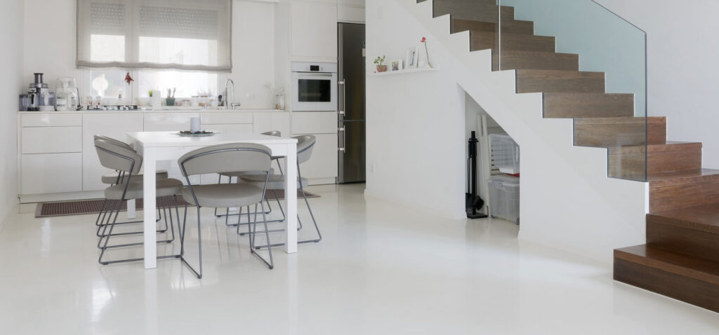 Spacious, modern kitchen with glossy white flooring reflects a minimalist design featuring a white dining table with gray chairs, sleek white cabinetry, and stainless-steel appliances. To the right, a staircase with wooden steps and glass balustrade ascends beside a clean white wall, enhancing the room's contemporary aesthetic.