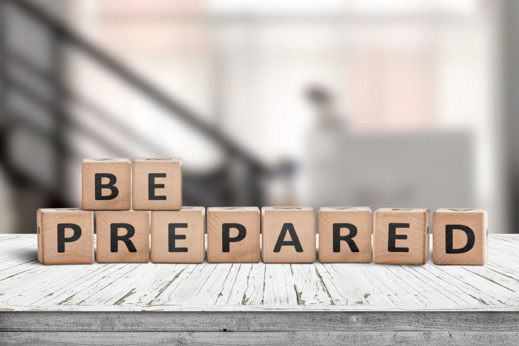 Wooden blocks with bold black letters spelling out the motivational message 'BE PREPARED' are arranged in a staggered line on a weathered white circular table. The blurred background features a staircase, contributing to the focused and encouraging atmosphere of the composition.