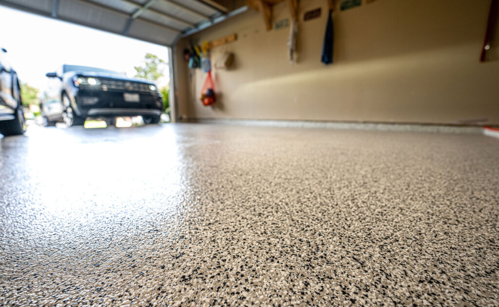 Close-up of a speckled epoxy garage floor with a blurred background showing a parked car and organized tools on the wall, illustrating a clean and maintained residential garage space.