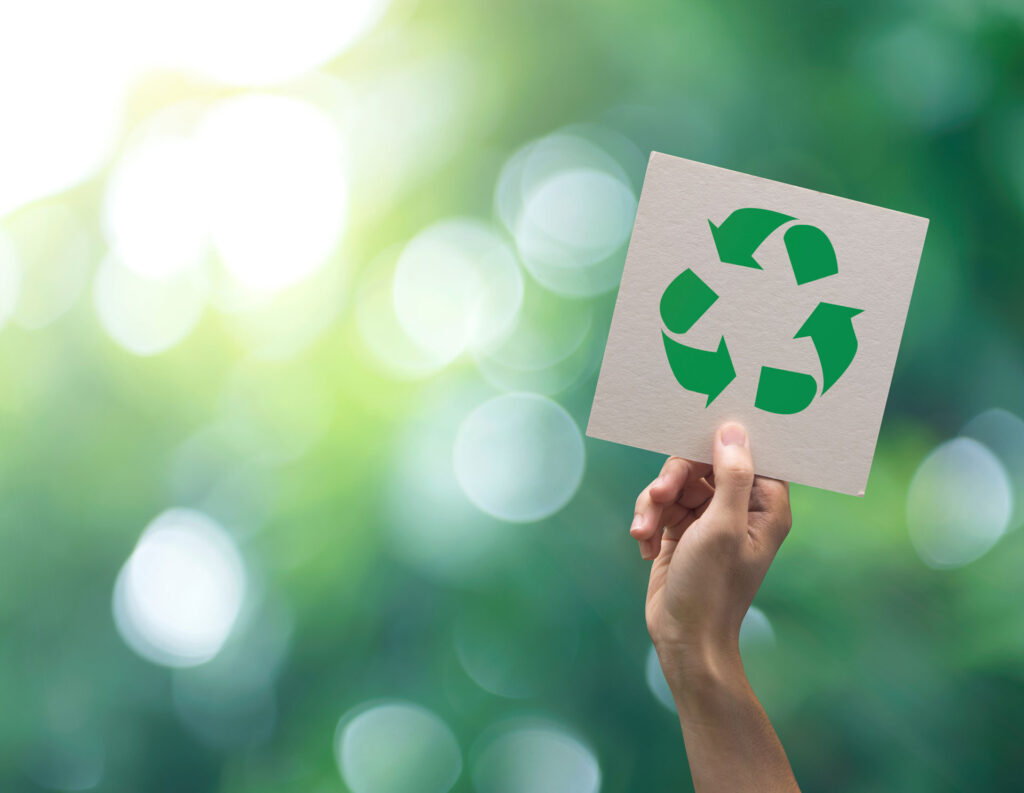 A hand holding up a card with the recycling symbol cut out, set against a bokeh background of lush green foliage illuminated by sunlight, symbolizing environmental awareness and the importance of recycling such things as concrete.