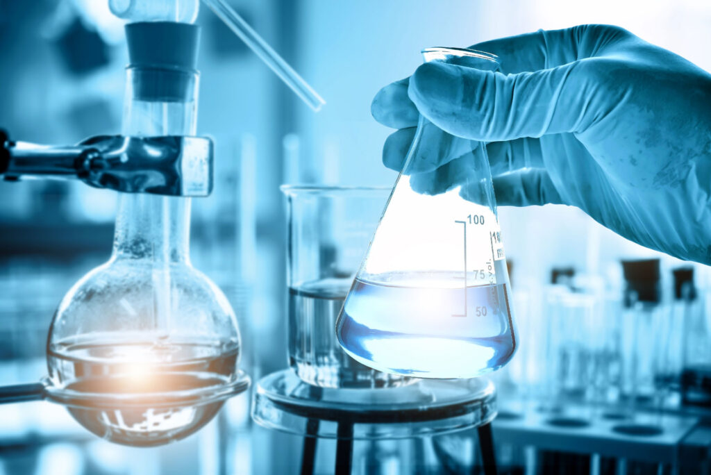 A gloved hand holding a flask with a blue liquid over a Bunsen burner in a laboratory setting, with other scientific glassware and a blurred background suggesting a research environment.