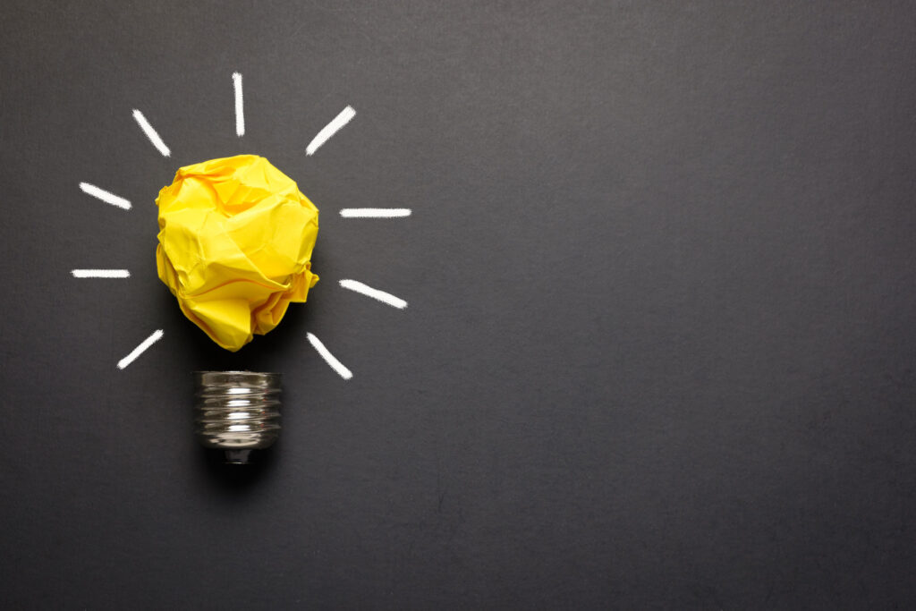 Creative representation of an idea with a lightbulb base and a crumpled yellow paper ball on top, drawn white lines mimic light rays on a dark gray background, conveying a concept of innovation or inspiration.