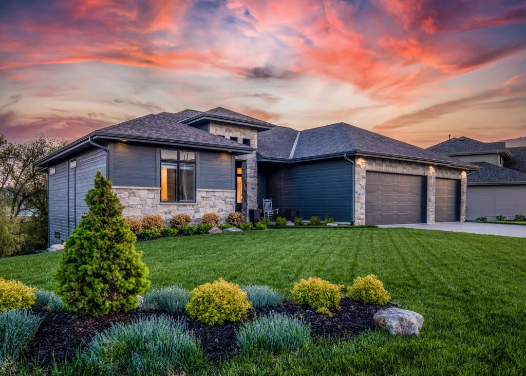 Modern single-story home with stone and dark siding exterior, featuring a double garage and well-manicured lawn with ornamental shrubs and a conifer tree. The house is set against a dramatic sunset sky with hues of pink and orange. A modern home perfect for aesthetic epoxy coatings inside and outside.