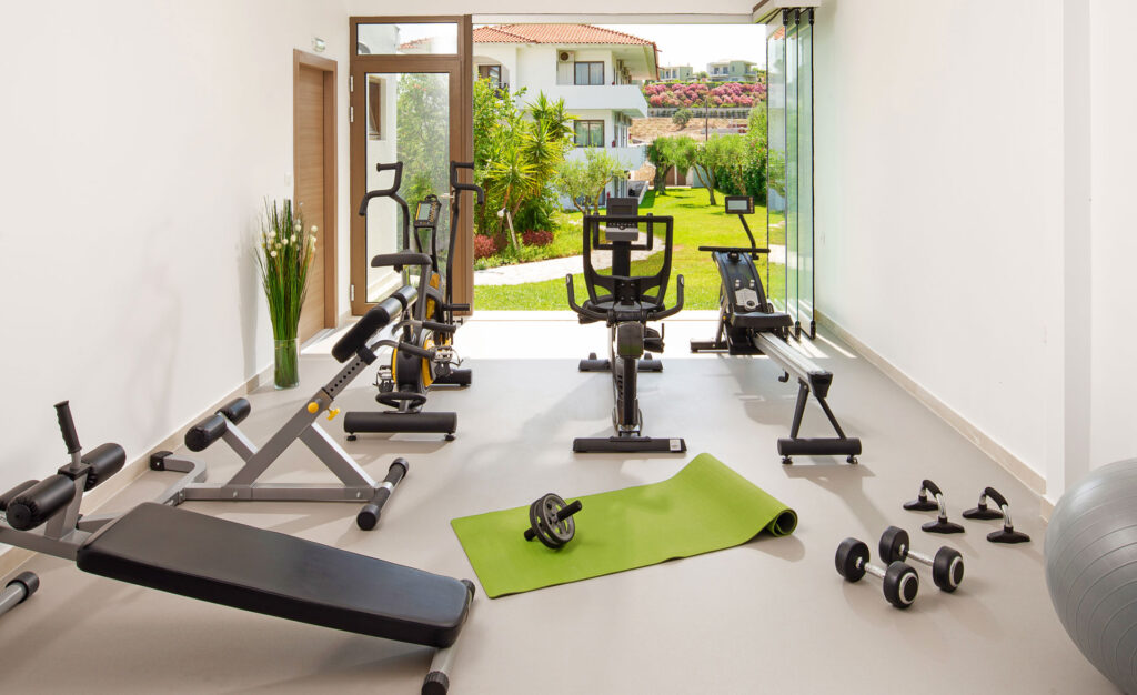 Home gym setup with various exercise equipment including a weight bench, stationary bikes, rowing machine, dumbbells, and a yoga mat on a bright, airy space with large glass doors opening to a green garden.