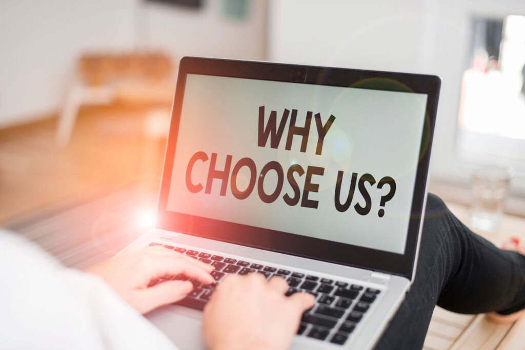 A person's hands are typing on a laptop with a screen displaying the message 'WHY CHOOSE US?' in bold letters. The background is bright, with sunlight streaming in, suggesting a work-from-home environment or a casual business setting while considering the benefits of a decision.