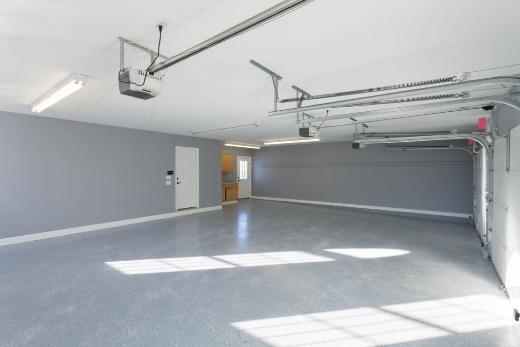 Spacious and clean residential garage interior with a newly applied grey epoxy floor and walls. The garage door's mechanical system is visible along with fluorescent lights on the ceiling. Natural light streams in through the window, casting soft shadows on the floor, creating a bright and organized space.