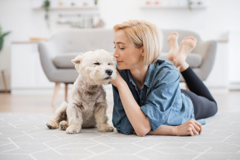 A serene moment as a woman with short blonde hair, wearing a casual denim shirt and black leggings, lies on her stomach on a patterned rug and gently touches noses with her fluffy white dog. The room is bright and airy, with a modern sofa in the background, depicting a peaceful, homey scene.