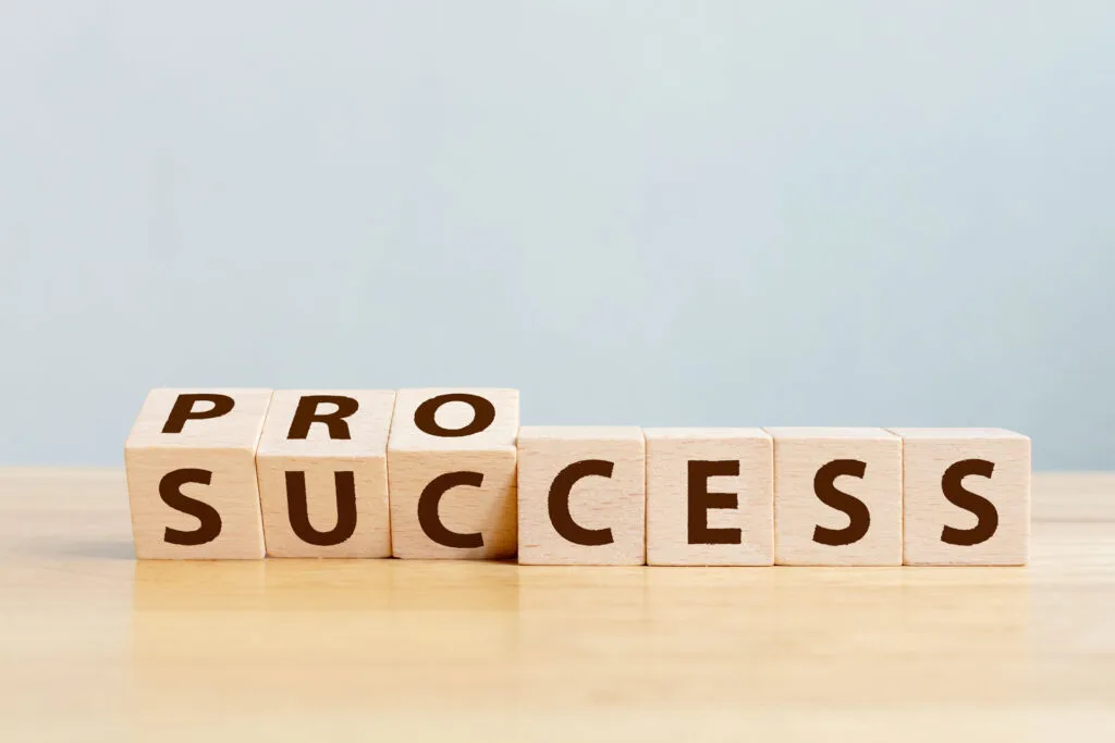 Wooden blocks with letters on a table spelling out 'PROCESS' with the last two blocks turned to reveal the word 'SUCCESS', symbolizing the idea that success is the result of a process. The backdrop is a soft blue, providing a calm and clear setting that complements the positive message.