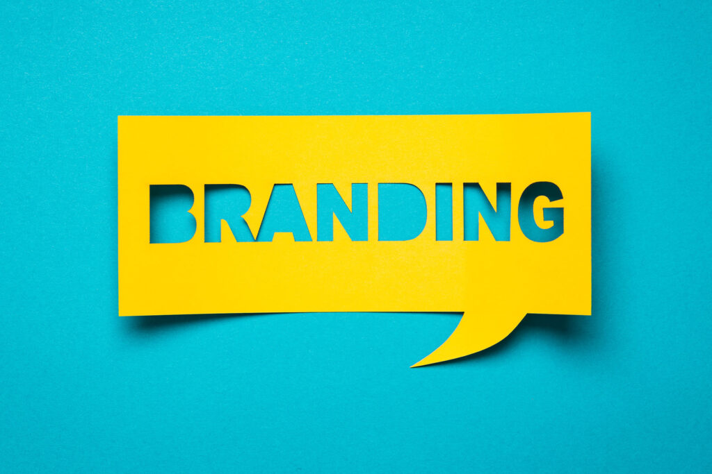 A vibrant yellow speech bubble cut-out with the word 'BRANDING' in bold, capitalized letters on a striking turquoise background. The design is modern and eye-catching, reflecting the concept of brand identity and marketing in a visually impactful way.