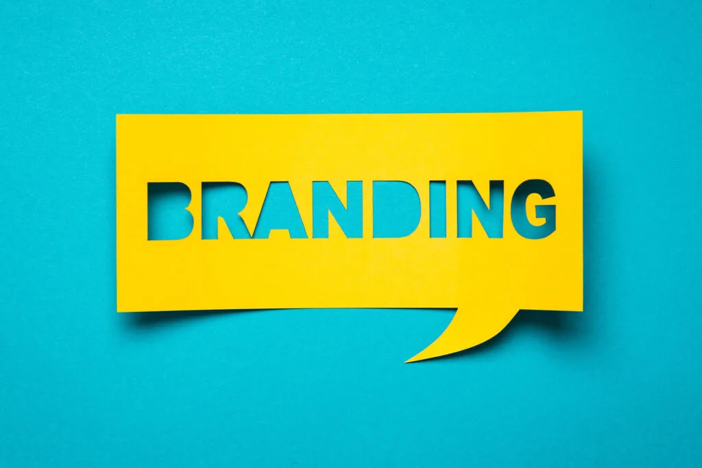 A vibrant yellow speech bubble cut-out with the word 'BRANDING' in bold, capitalized letters on a striking turquoise background. The design is modern and eye-catching, reflecting the concept of brand identity and marketing in a visually impactful way.