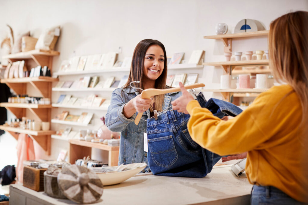 Inside a bright boutique, a smiling sales assistant in a denim jacket hands a pair of jeans to a customer. The store is filled with neatly organized artisanal goods on wooden shelving, creating a warm and inviting shopping atmosphere. The exchange highlights a personal and attentive retail experience.