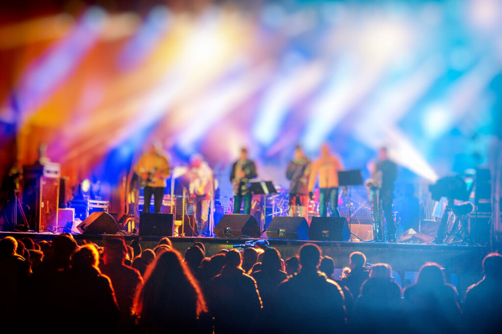 Vibrant and colorful blurred scene of a live concert with silhouettes of a crowd in the foreground and the band on stage in the background. The bright stage lights cast dynamic beams across the venue, creating a lively and energetic atmosphere typical of musical performances.