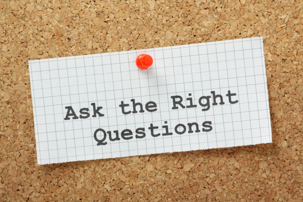 A piece of white graph paper pinned to a corkboard with a red pushpin, with the phrase 'Ask the Right Questions' typed in bold, black letters. The simple yet meaningful message stands out against the textured beige background of the cork, suggesting the importance of critical thinking and inquiry in various contexts.