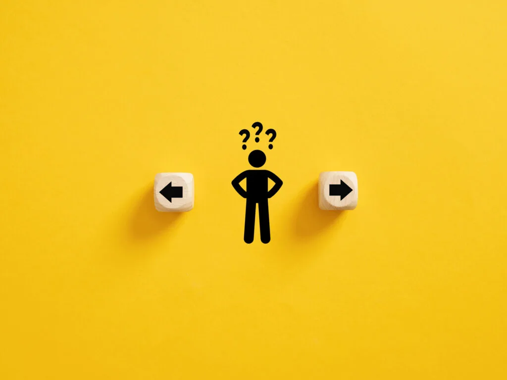 A conceptual image on a bright yellow background showcasing decision-making, with a stick figure standing between two wooden cubes, each pointing in opposite directions with black arrows. Above the figure are question marks, symbolizing the individual's indecision or the process of choosing between two different paths or options such as epoxy coatings or epoxy paint as flooring solutions.