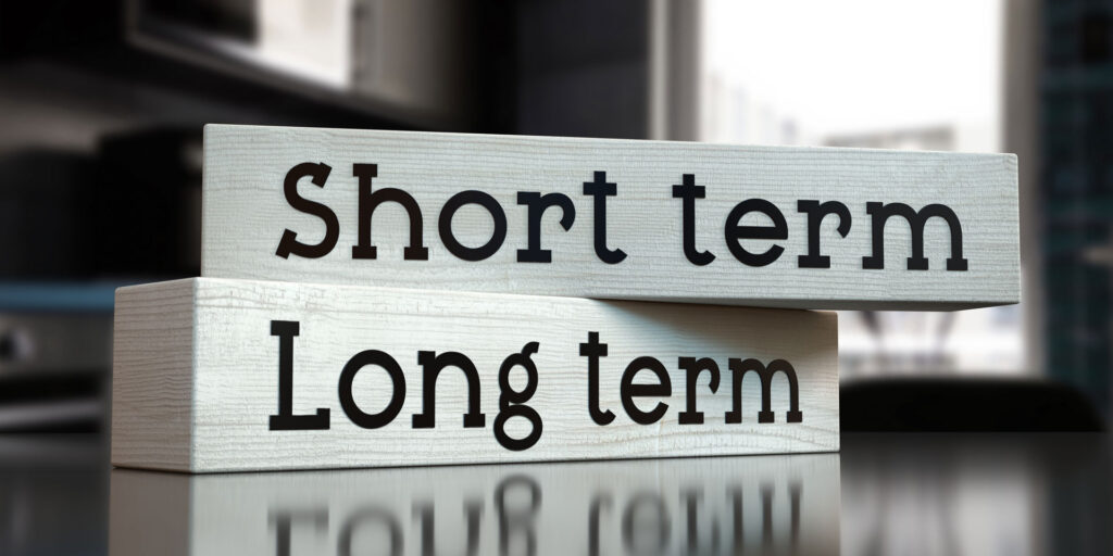 Two wooden blocks on a reflective surface with the words 'Short term' and 'Long term' printed in bold black letters. The 'Short term' block is stacked on top of the 'Long term' block, illustrating a conceptual juxtaposition often encountered in planning, strategy, and decision-making contexts such as the longevity and durability of flooring types.
