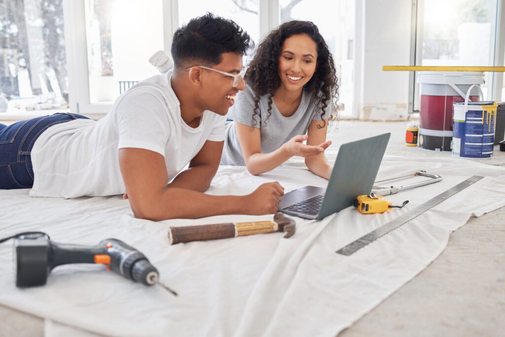 A cheerful young couple engaging in a home renovation project. The man, wearing a white t-shirt and glasses, and the woman, in a gray top, are lying on a drop cloth and looking at a laptop screen together. Various DIY tools, including a hammer, tape measure, and drill, are scattered around them, with paint cans and a ladder in the background, indicating an active modern aesthetics home improvement setting.