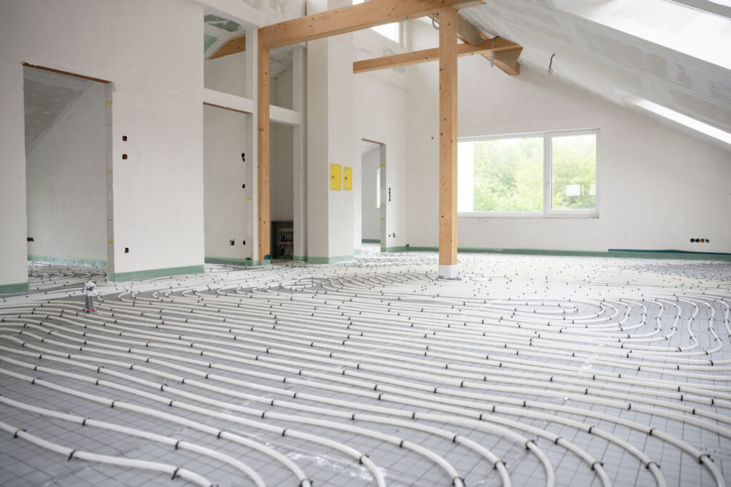 An expansive view of a room under construction with a radiant floor heating system installation in progress. The network of white pipes is methodically arranged across the floor, leading to junction boxes on the walls. Wooden beams support the ceiling, and natural light streams in through the windows, illuminating the space and highlighting the precise layout of the heating system prior to the application of the epoxy coating.