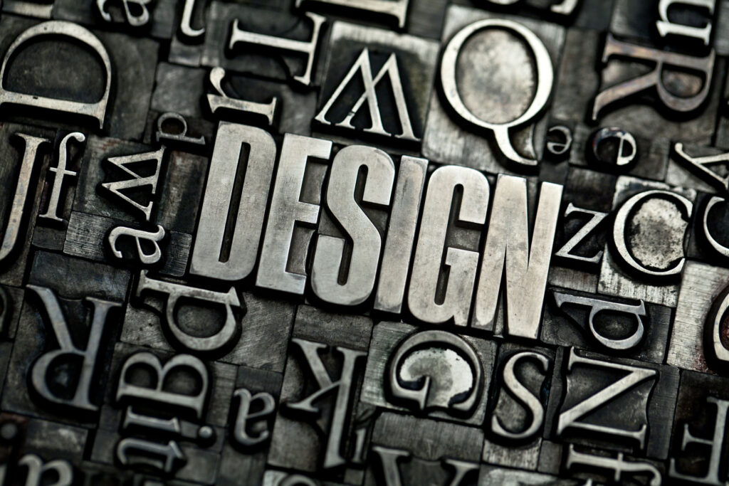 A textured assortment of metallic letterpress type blocks, with the word 'DESIGN' in bold and in focus at the center. Surrounding it are various letters and punctuation marks in different fonts and sizes, creating a monochromatic mosaic that conveys a vintage and artistic atmosphere associated with typography and graphic design or creativity.