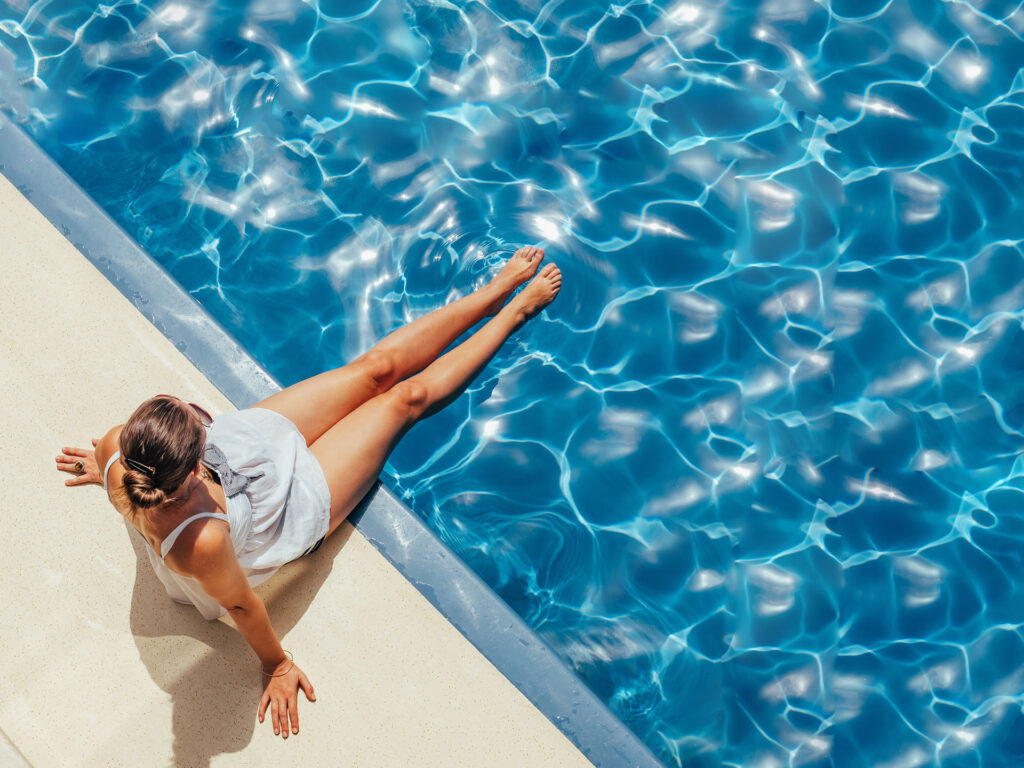 Overhead view of a person lounging on an epoxied pool deck by the edge of a sparkling pool, with their legs dipped in the water. The individual is wearing a white sleeveless top and has their hair tied back, enjoying the sun on a clear day. The water's surface is a vibrant blue with light patterns dancing across it, suggesting a peaceful and leisurely atmosphere.