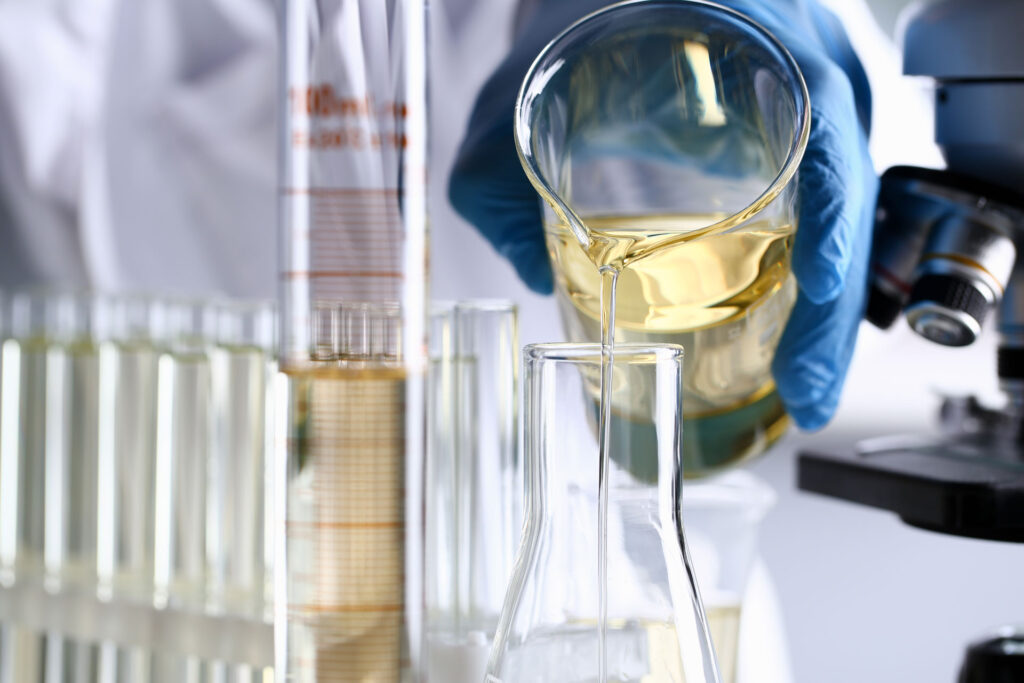 A close-up image capturing the precise moment a clear yellow liquid is being poured from a beaker into a test tube by a person wearing blue protective gloves in a laboratory setting. In the background, several other test tubes are lined up next to a graduated cylinder, with a microscope partially visible, indicating an active scientific research environment.