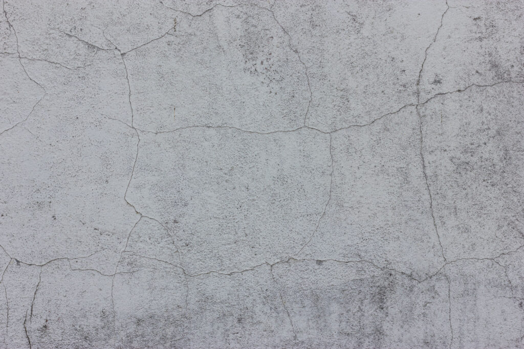A close-up of a gray concrete surface with a textured finish and a network of fine cracks throughout, displaying the natural wear and tear of the material.