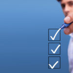 Close-up of a person in a white shirt ticking off a checklist with a blue pen, against a blurred blue background. The checklist contains three checkboxes, with the top two already marked with a check, indicating completed tasks.