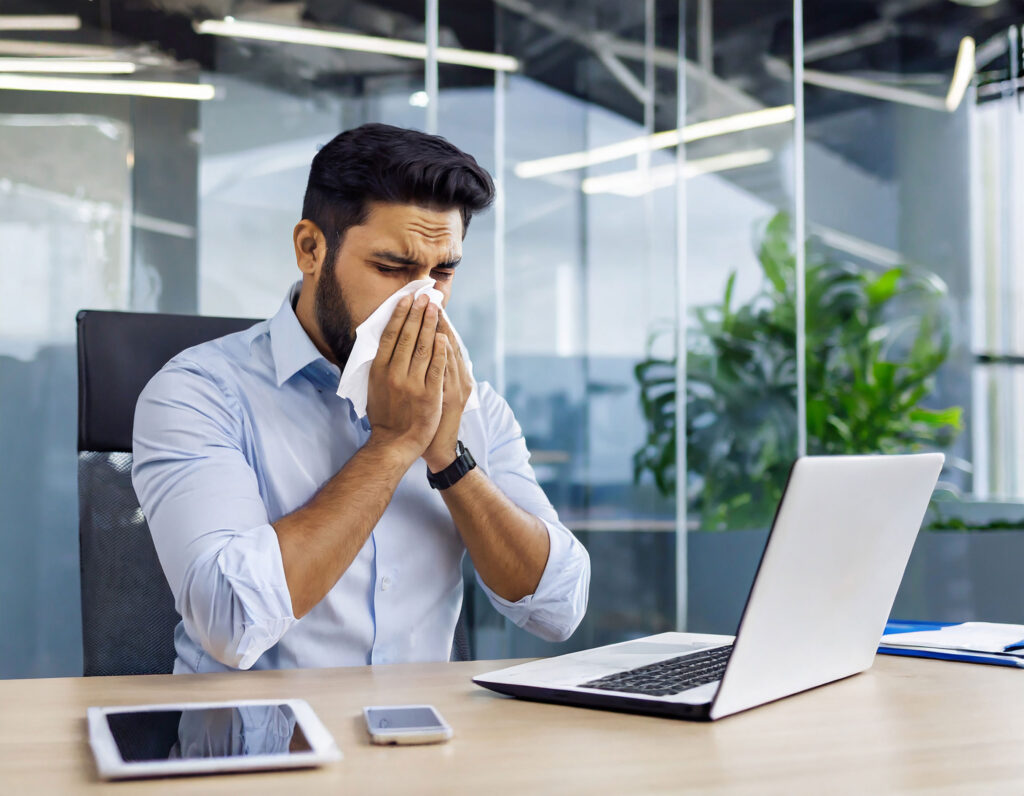 Business man suffering from allergies is blowing his nose while sitting in his office at a desk with an ipad, phone and laptop computer. He is wearing a light blue dress shirt with the sleeves rolled up. There are glass walls separating the offices and a large green plant in the background. Epoxy flooring can help relieve his allergies compared to other flooring choices.