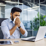 Business man suffering from allergies is blowing his nose while sitting in his office at a desk with an ipad, phone and laptop computer. He is wearing a light blue dress shirt with the sleeves rolled up. There are glass walls separating the offices and a large green plant in the background. Epoxy flooring can help relieve his allergies compared to other flooring choices.