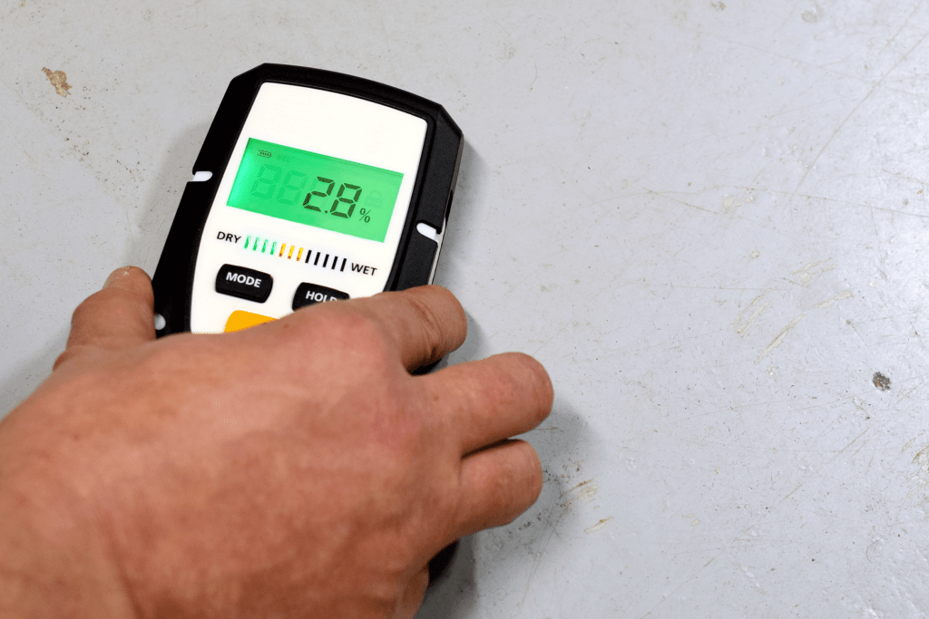 Man's hand holding a moisture meter against a concrete floor to determine the level of moisture in the flooring. This helps to determine if moisture mitigation is needed prior to installing an epoxy flooring.