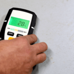 Man's hand holding a moisture meter against a concrete floor to determine the level of moisture in the flooring. This helps to determine if moisture mitigation is needed prior to installing an epoxy flooring.